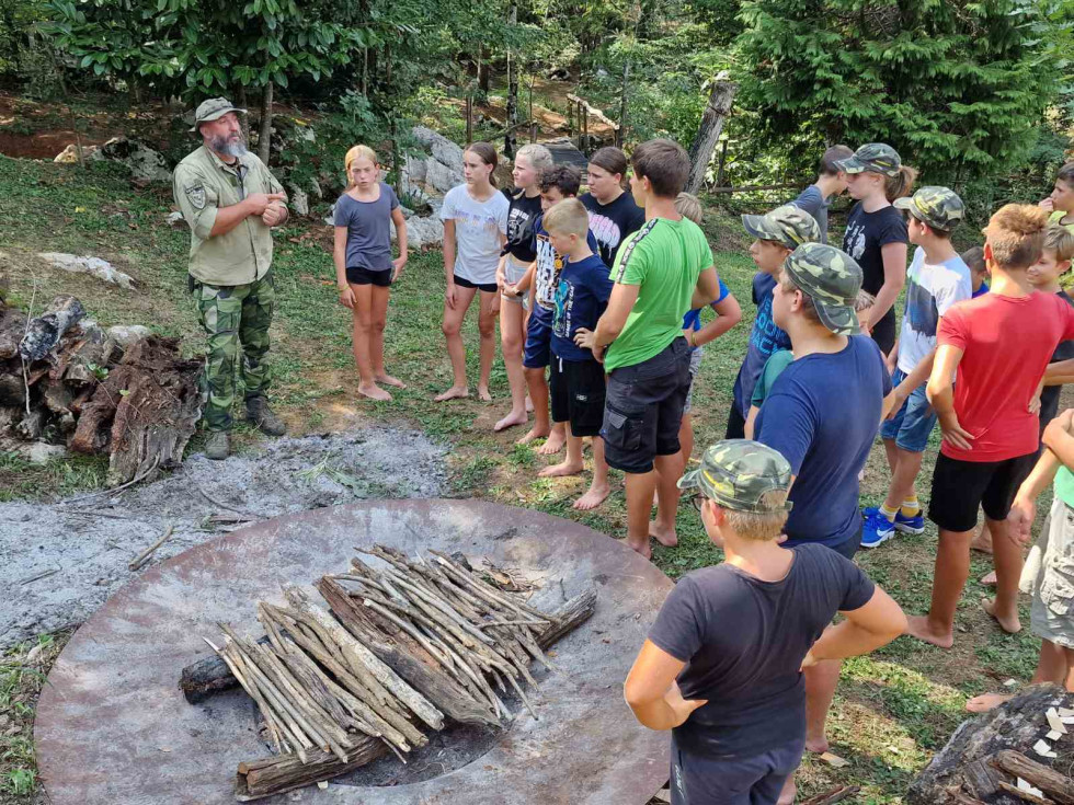 Otroci, udeleženci tabora stojijo okrog lesa za ogenj in poslušajo inštruktorja
