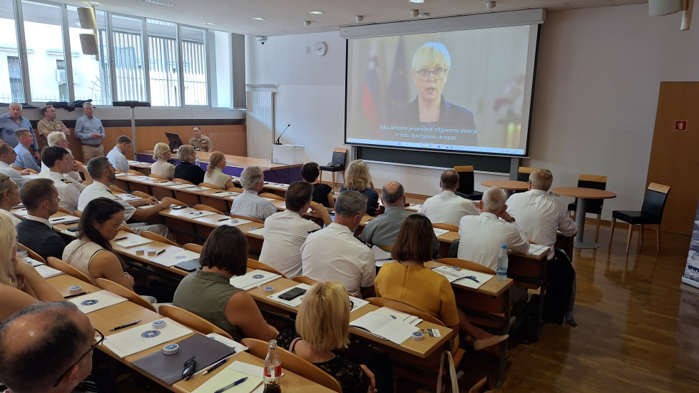 Udeleženci konference sedijo v avditorju in spremljajo pozdravni nagovor predsednice republike na velikem zaslonu pred njimi.