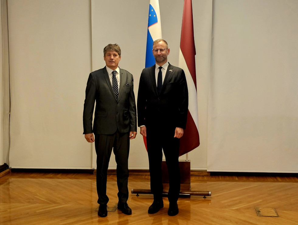 Minister in sekretar stojita pred zastavama Slovenije in Latvije