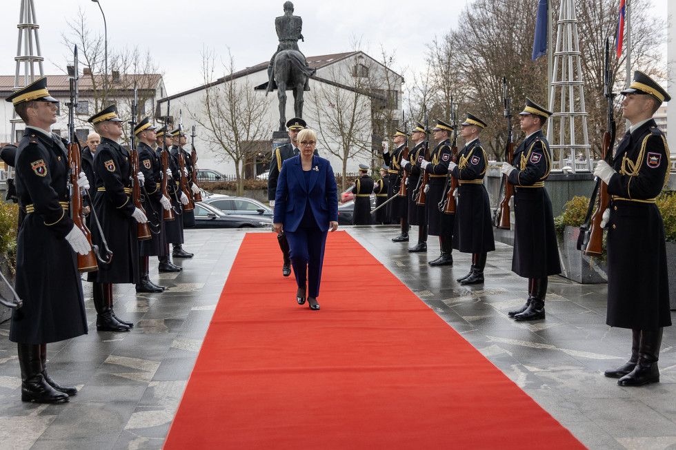 Predsednica hodi po preprogi med postrojenimi gardisti. V ozadju je kip generala Maistra