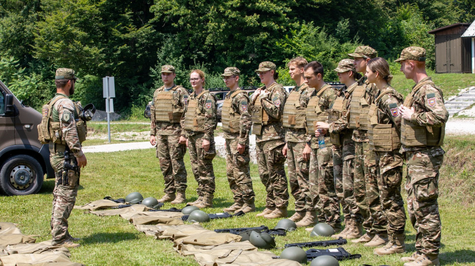 Udeleženci tabora v uniformah Slovenske vojske med usposabljanjem stojijo na vadišču. Pred njimi je inštruktor.