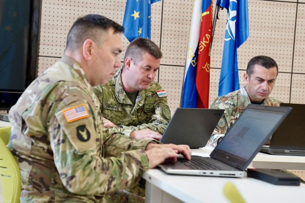 Participants sit at their computers. Behind them are the flags of the participating countries
