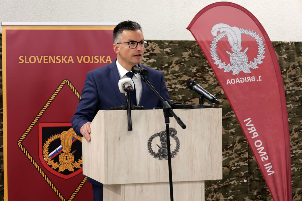 Minister med govorom stoji za govornico. Ob njem je rdeč logotip prve brigade