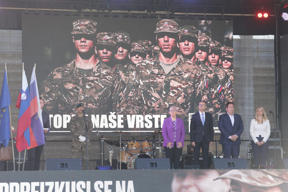 Predsednica, minister in drugi gostje stojijo na odru med slovesnostjo. Za njimi je projekcija s sliko vojakov
