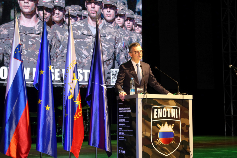 Minister Šarec stoji za govorniškim pultom na katerem je grb Slovenske vojske z napisom: Enotni, na levi zastave in v ozadju slika postroja vojakov