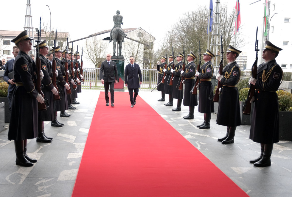 Ministra hodita po rdeči preprogi med postrojenimi gardisti. Za njima je spomenik generalu Maistru