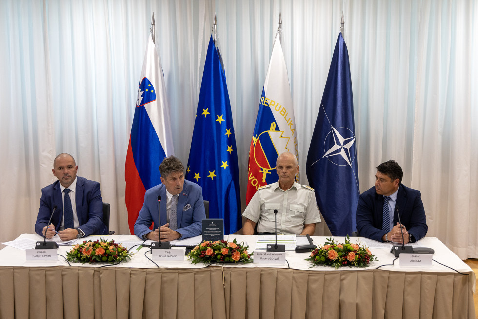 Udeleženci konference sedijo za mizo. Za njimi so zastave Slovenije, EU, Nato in Slovenske vojske