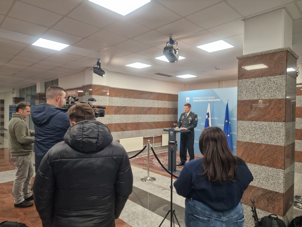Tiskovni predstavnik Slovenske vojske med izjavo stoji za govornico. Pred njim so snemalci, za njim pa moder pano ministrstva