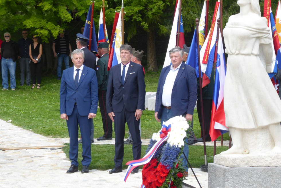 Minister in drugi visoki gostje stojijo pred spomeniki v parku. Za njimi so praporščaki z zastavami