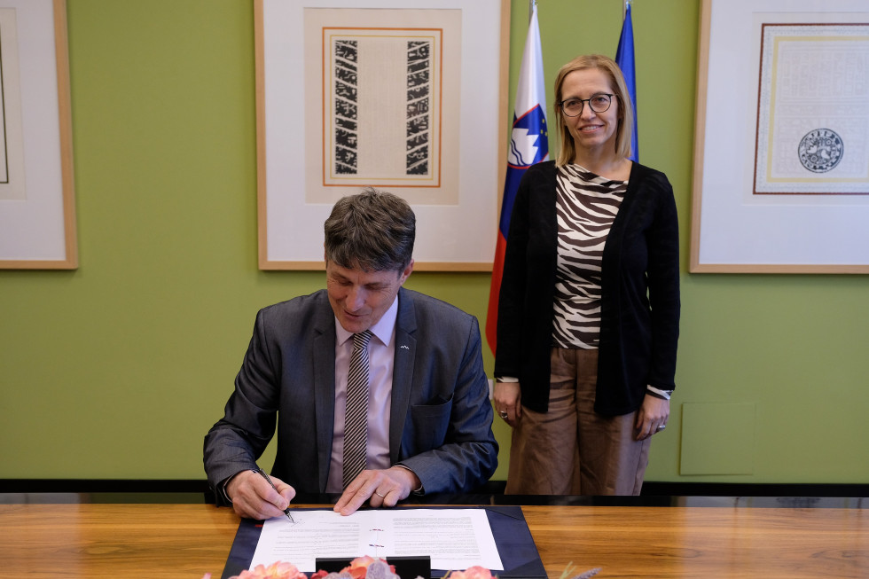 Minister za obrambo Borut Sajovic sedi za protokolarno mizo in podpisuje listino, ob njem na desni strani stoji ministrica za zdravje Valentina Prevolnik Rupel