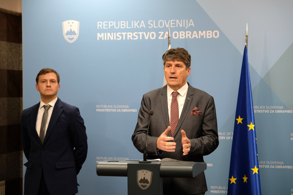 Minister med izjavo stoji z govornico. Za njim stoji direktor, pred modrim panojem in zastavama Slovenije in EU