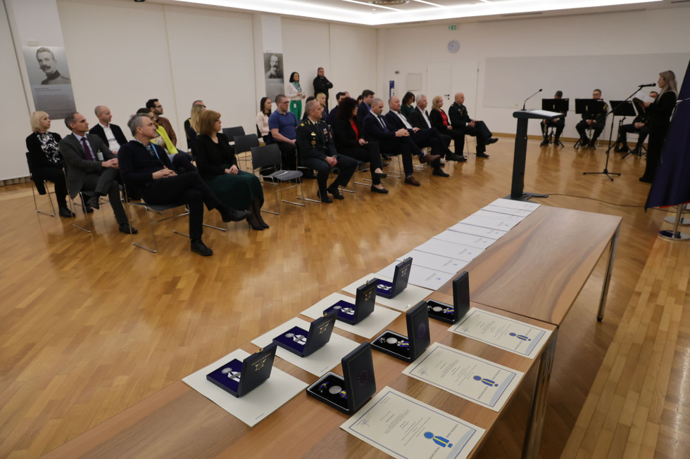 Udeleženci slovesnosti sedijo v dvorani. Pred njimi je govornica in mize z odlikovanji