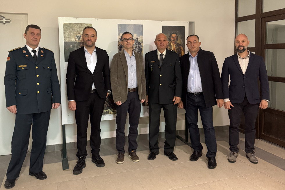 Šest udeležencev bilateralnega srečanja, štirje v civilnih oblekah in dva v vojaških uniformah, stoji pred panojem s tremi plakati iz črnogorske zgodovine. 