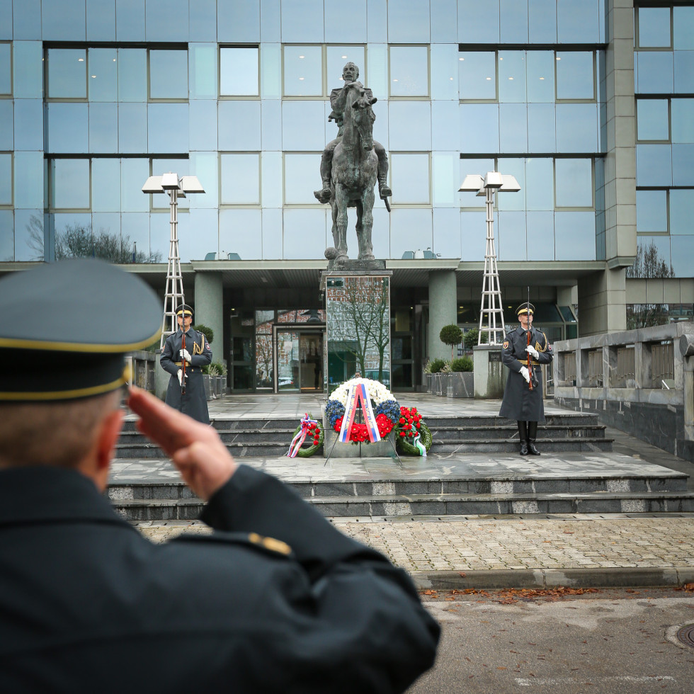 Gardist z roko v pozdravu. V ozadju je spomenik generalu Maistru pred stavbo ministrstva