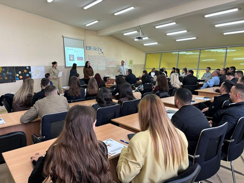 Udeleženci predavanja sedijo za mizami v predavalnici. V ozadju je predavatelj in zaslon s projekcijo