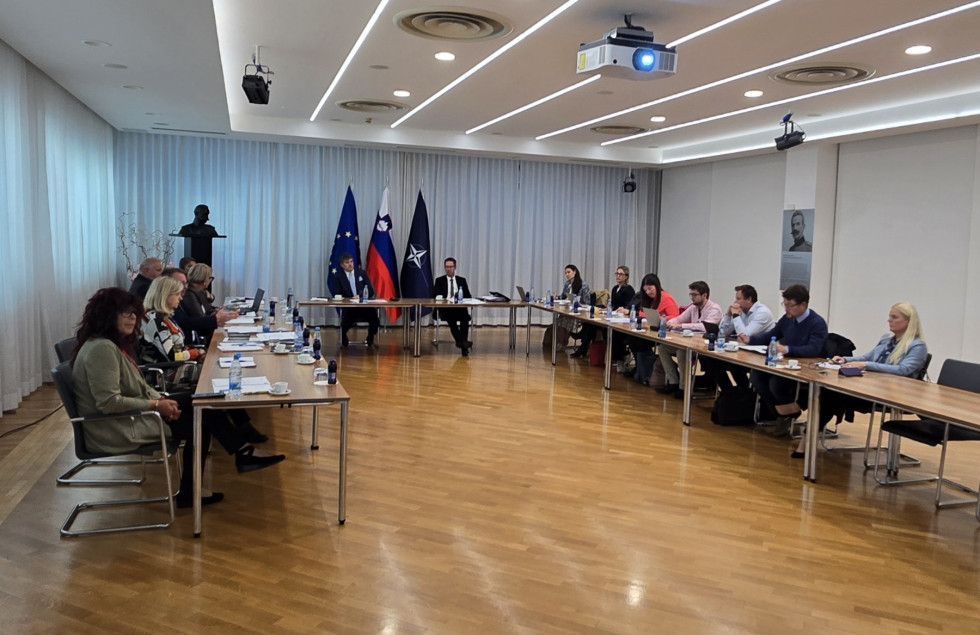Udeleženci sestanka sedijo za mizami postavljenimi v veliko dvorano