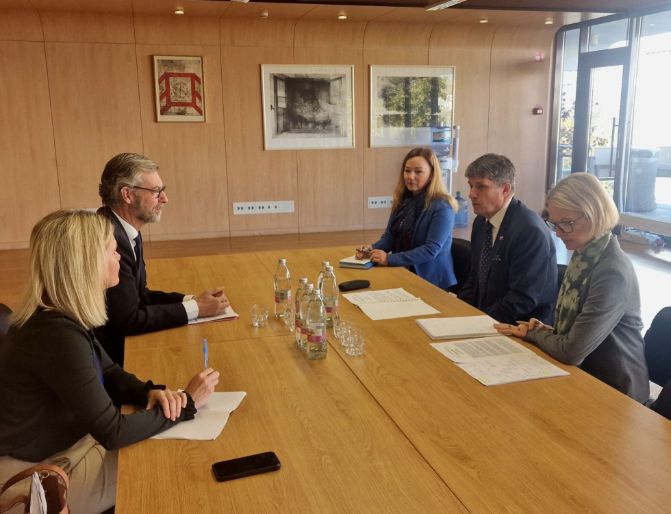 Minister in podpredsednik ter člani delegacij med pogovori sedijo za sejno mizo. V ozadju na steni sta umetniški sliki.