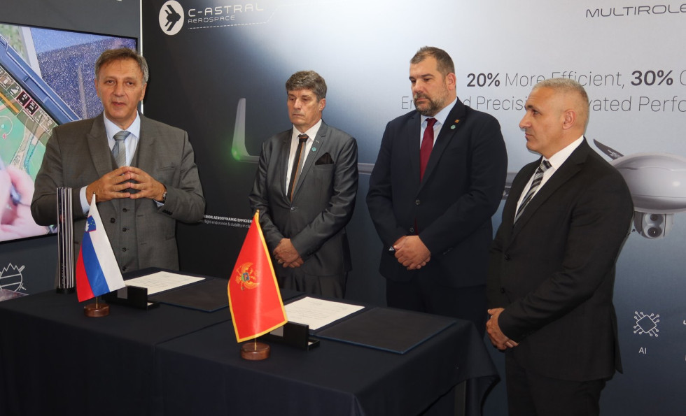 Direktorja in ministra za obrambo Slovenije in Črne gore stojijo za podpisnim pultom, na katerem sta postavljeni slovenska in črnogorska zastavica ter dokumenti za podpis. V ozadju je delno viden panel z informacijami o sistemu Belin.