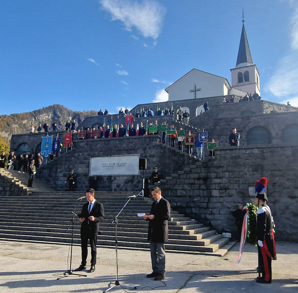 Državni sekretar Lovše in prevajalec stojita pred kamnitim stopniščem, ki vodi do kostnice in po katerem so razporejeni nosilci praporov udeležencev in delegacij ter častna straža. Na vrhu je viden del cerkve z zvonikom.