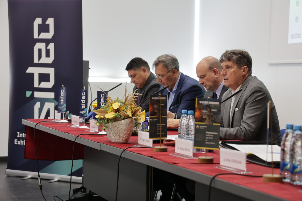 Udeleženci novinarske konference sedijo za mizo z mikrofoni in zastavicami z logotipi