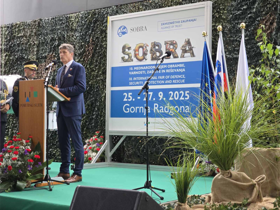 Minister med govorom stoji za govornico na odru. Za njim je pano z logotipom sejma.