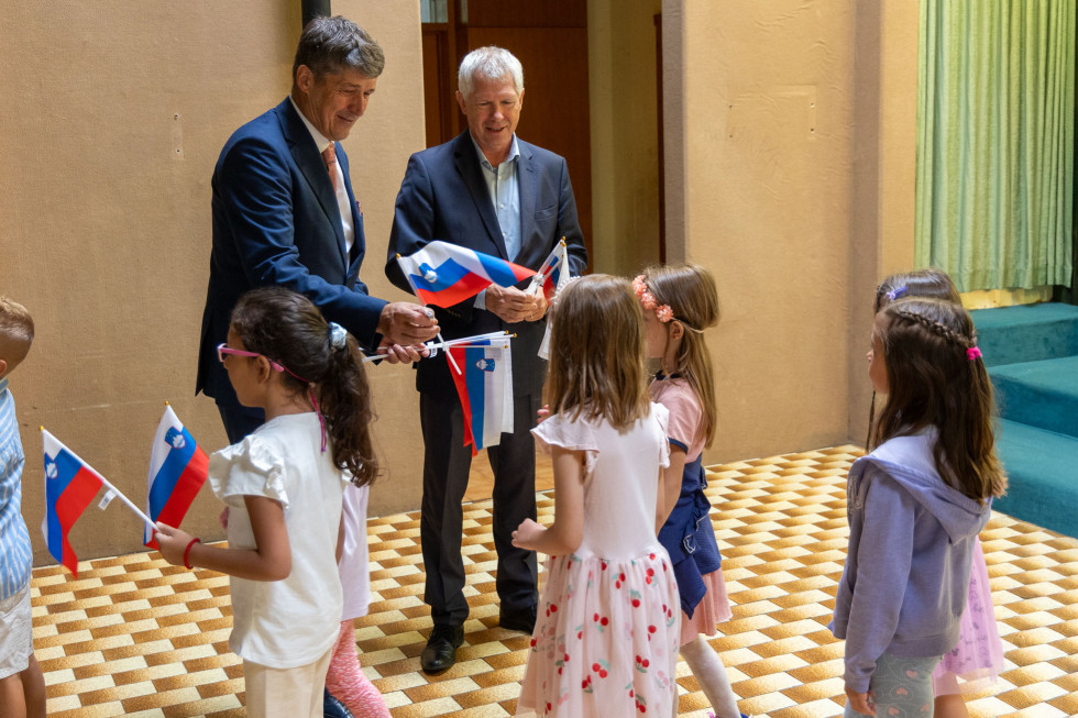 Minister Sajovic stoji pred nekaj otroki, ki čakajo, da jim minister podeli slovenske zastavice. 