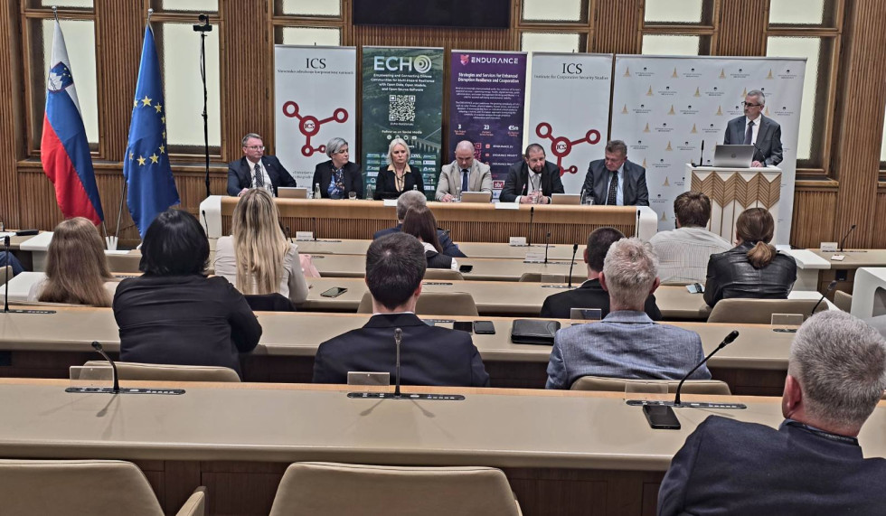 Dvorana državnega sveta s klopmi v katerih sedijo udeleženci. Nastopajoči v ozadju sedijo za mizo. Z njimi je platno s projekcijo in logotipi organizatorjev 