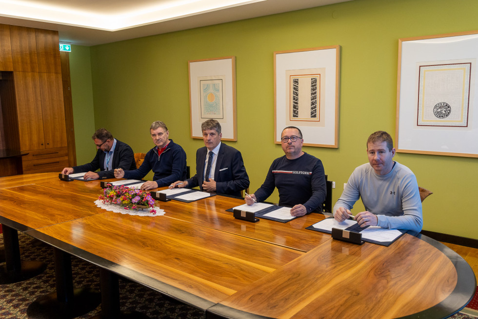 Podpisniki pogodbe sedijo za dolgo mizo. Za njimi na steni so umetniške slike