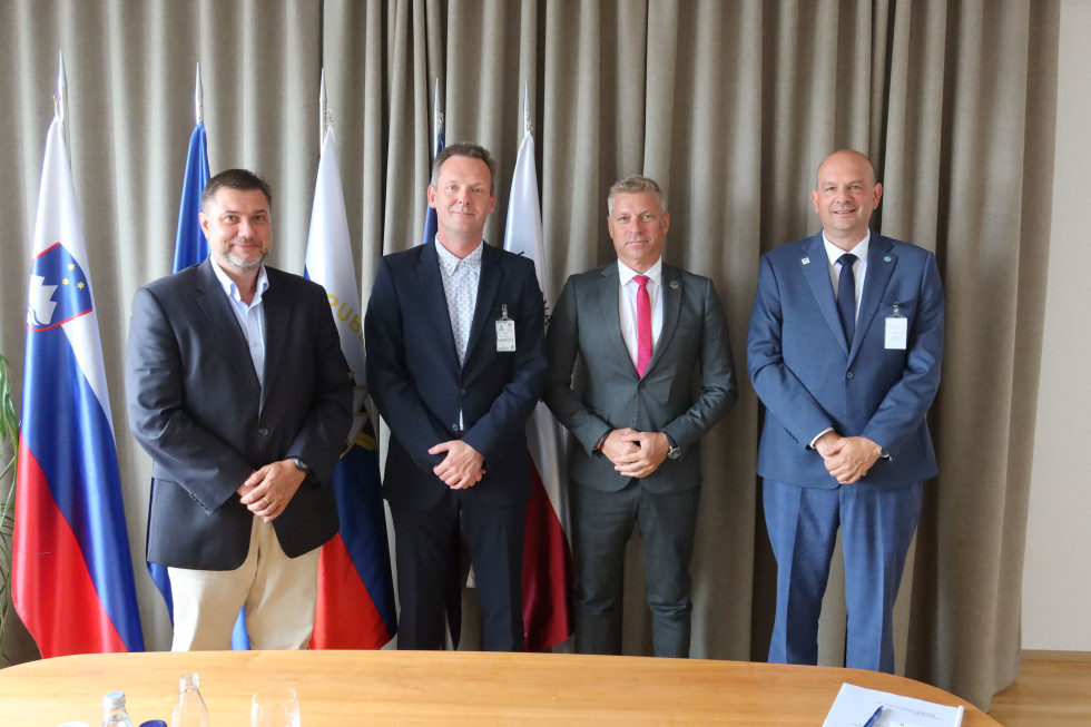 Skupinska fotografija ministra, njegovega sodelavca in obeh predstavnikov GOIS, ki stojijo drug poleg drugega. V ozadju je pet praporov z zastavami in stena s sivo zaveso.