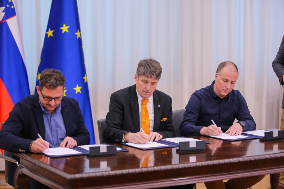 Minister in predstavnika sindikata ter stavkovnega odbora sede za mizo podpisujejo sporazum. Za njimi sta zastavi Slovenije in EU