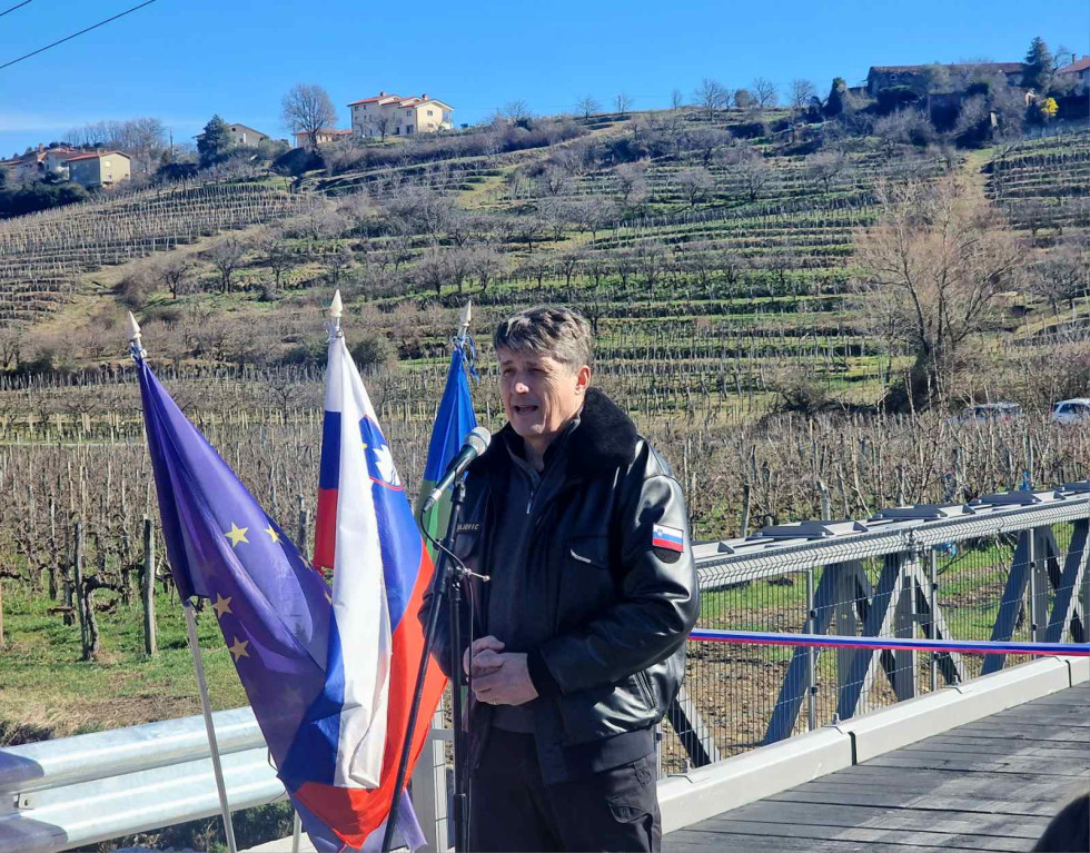 Minister med otvoritvenim nagovorom stoji pred mostom. Za njim so zastave Slovenije, EU in občine Brda