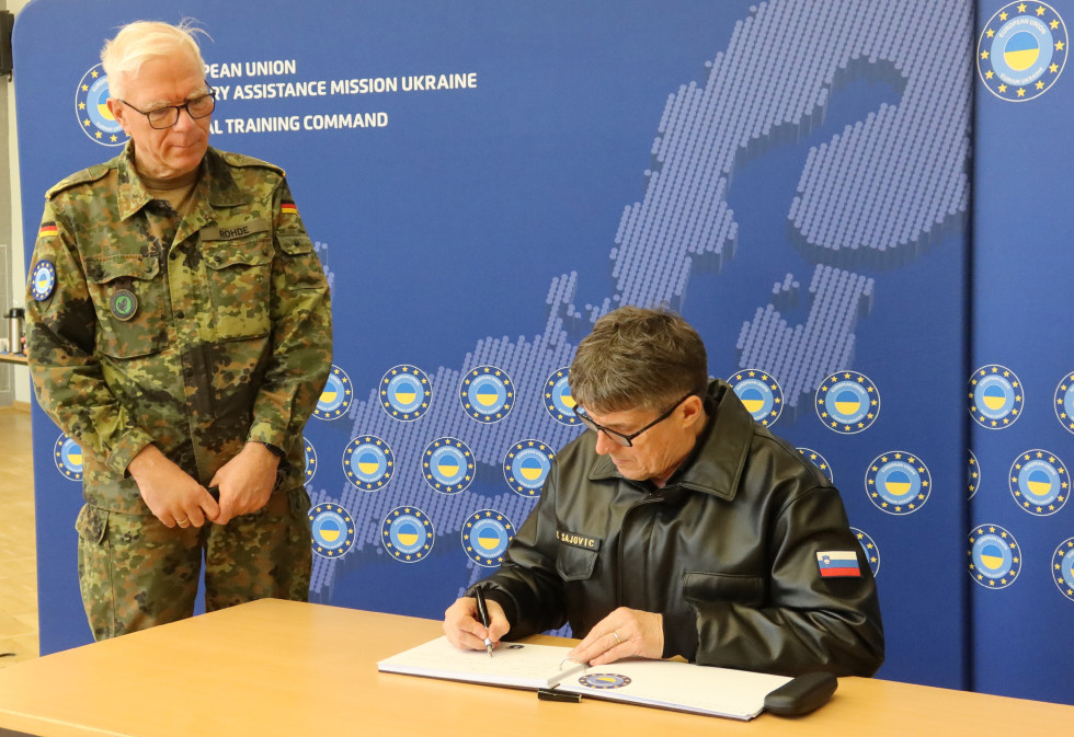 Minister se vpisuje v knjigo gostov. Ob njem stoji gostitelj, za njima pa je moder pano