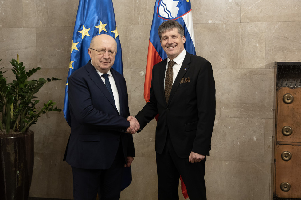 Komisar Kolbilius in minister Sajovic stojita in se rokujeta pred evropsko in slovensko zastavo.