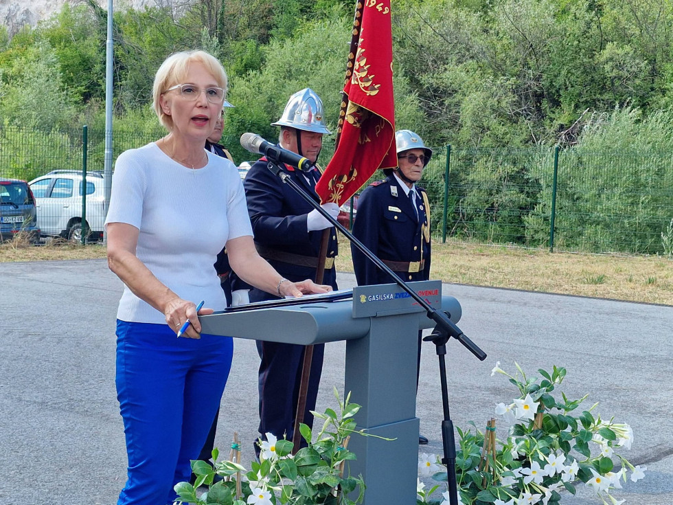 Sekretarka med nagovorom stoji za govornico na poligonu. Za njo sta gasilca z zastavo društva  