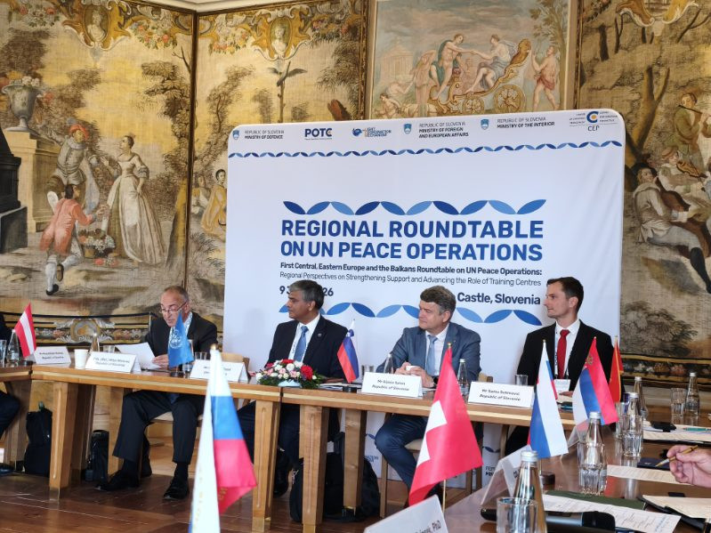Moderator in trije uvodni razpravljavci sedijo za konferenčno mizo v grajski dvorani. Za njimi je pano z belo podlago in modrim logotipom konference. Na steni v ozadju so tapiserije z motivi življenja na gradu iz obdobja baroka.