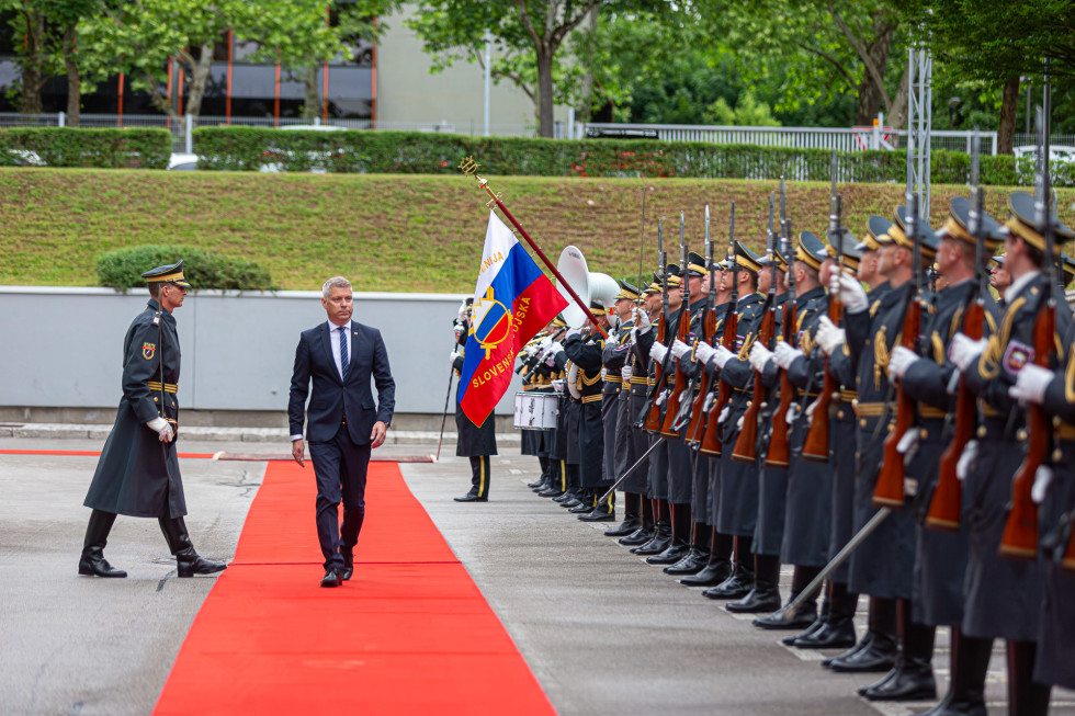Minister Hajdinjak hodi po rdeči preprogi mimo orkestra in postroja častne čete Slovenske vojske, eden od gardistov drži prapor s slovensko zastavo, poleg njega na levi stoji poveljujoči častni četi.
