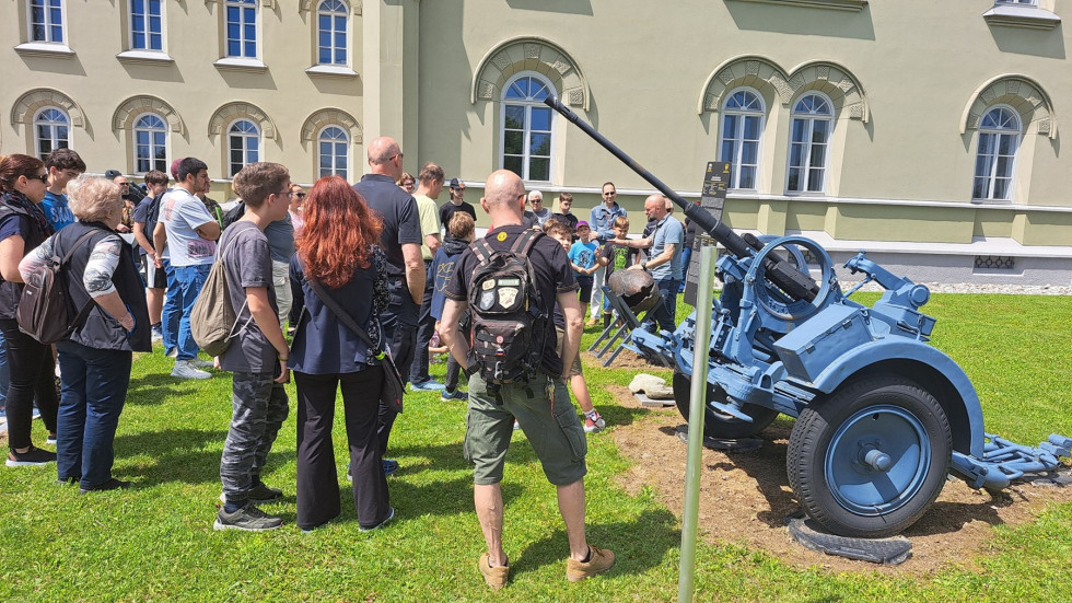 Skupina ljudi stoji v polkrogu na zelenici pred vojaškim objektom in si ogleduje razstavljene vojaške eksponate.