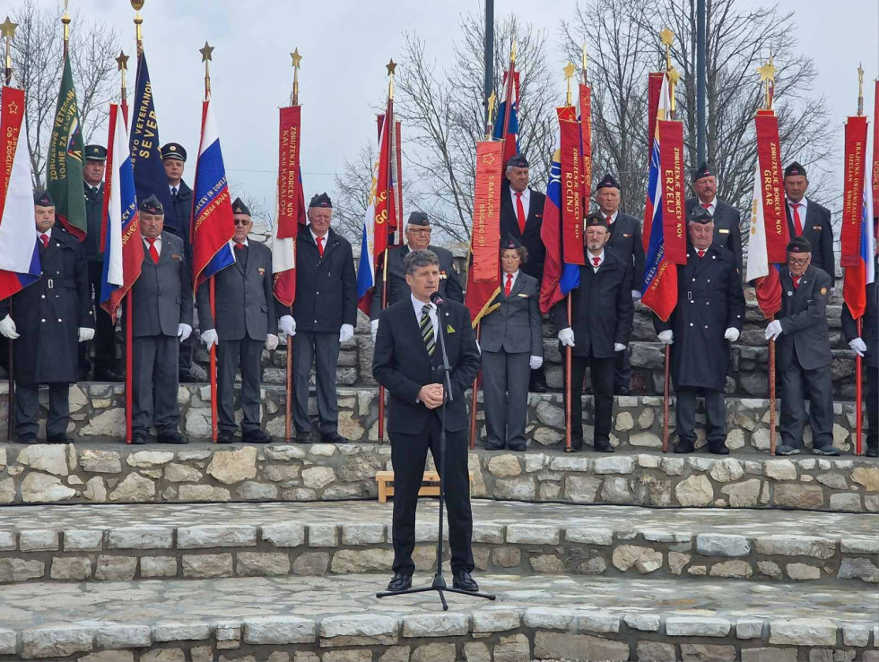 Minister med govorom stoji na kamnitih tleh. Za njim so razporejeni nosilci praporjev.