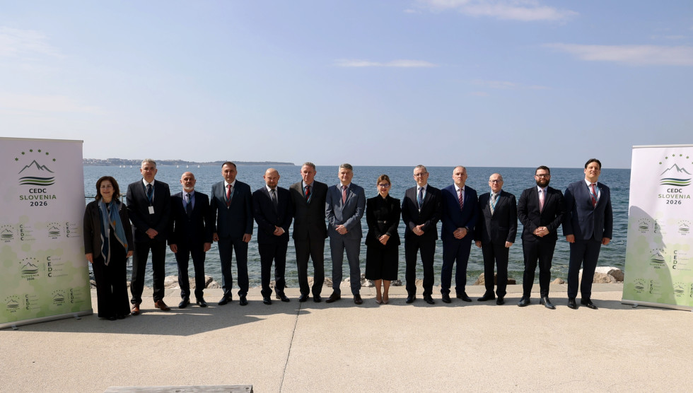 Skupinska fotografija udeležencev srečanja na obali v Portoržu. Udeleženci stojijo v vrsti, na levi in desni sta panoja z logom CEDC, v ozadju je morje.