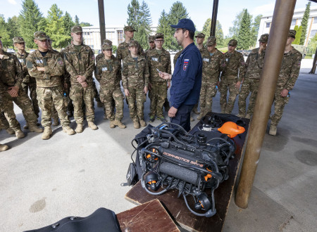 Udeleženci tabora oblečeni v vojaške uniforme stojijo v polkrogu pod nadstreškom in poslušajo mornariškega inštruktorja Slovenske vojske v modri uniformi, ki stoji pred skupino.