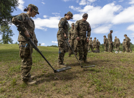 Udeleženki tabora oblečeni v vojaški uniformi na poligonu vadita z detektorjem kovin. Zraven stoji pripadnik Slovenske vojske, ki ju inštruira.