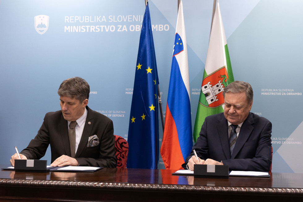 Minister in župan sede za mizo podpisujeta sporazum. Za njima so zastave Slovenije, EU in Ljubljane ter moder pano ministrstva