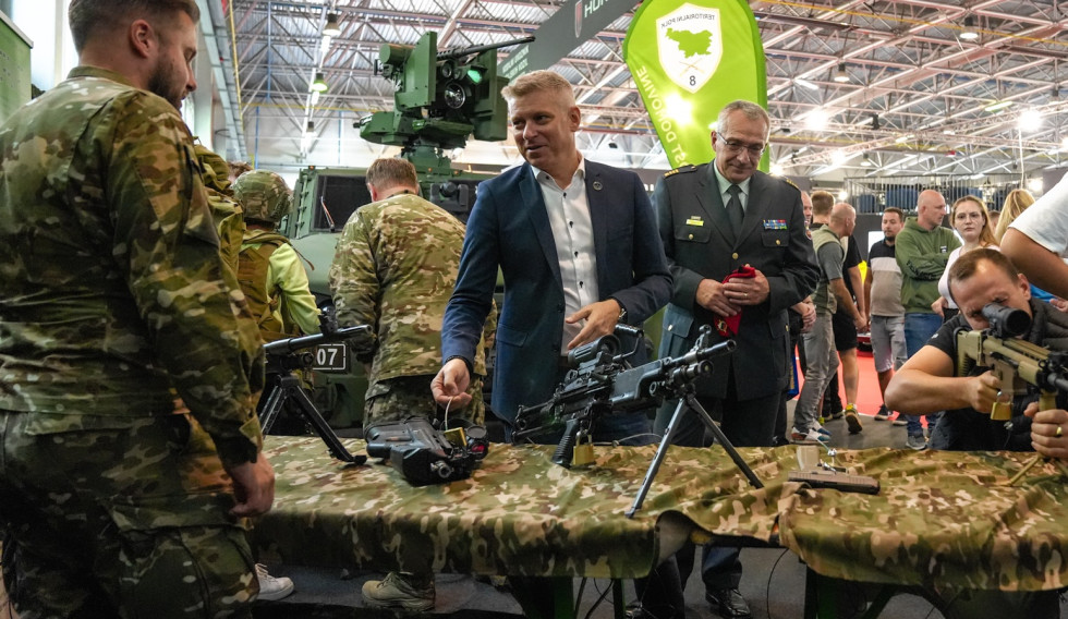 Minister stoji pred razstavno stojnico Slovenske vojske, si ogleduje razstavljeno oborožitev in se pogovarja z vojakom, ki stoji v razstavnem prostoru. V ozadju so obiskovalci in vojaško oklepno vozilo.