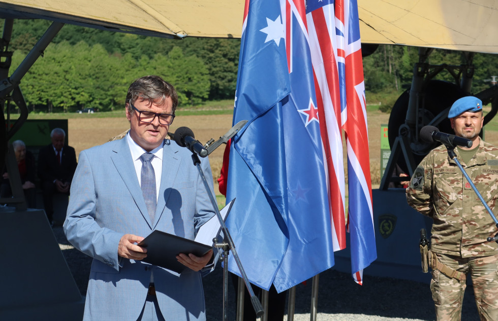 Sekretar med govorom stoji pred mikrofonom na stojalu. Za njim so zastave zavezniških držav med drugo svetovno vojno