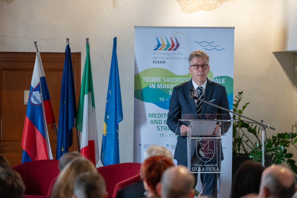 Državni sekretar Lovšin stoji za govornico, na levi zastave