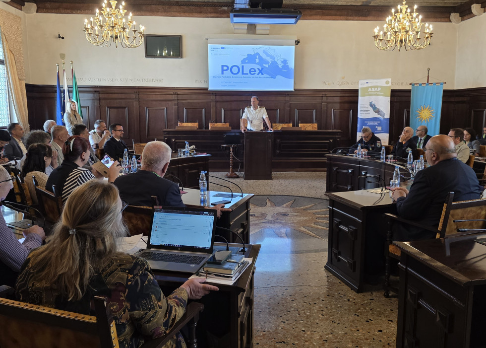 Zbrani v veliki sejni sobi poslušajo predavatelja, za njim projekcija