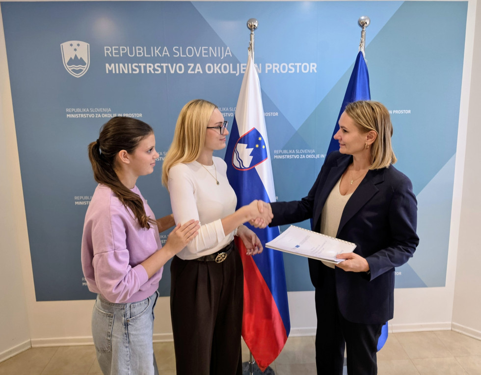 Ministrica Rifelj prevzema gradivo od predstavnic mladih, v ozadju zastavi in napis Ministrstvo za okolje in prostor
