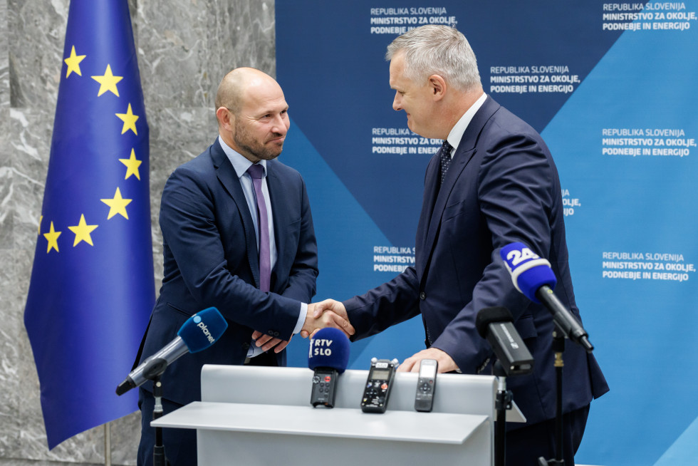 Dve osebi v poslovnih oblekah si rokujeta pred govorniškim pultom. V ozadju sta zastavi Slovenije in Evropske unije ter moder pano z napisom.
