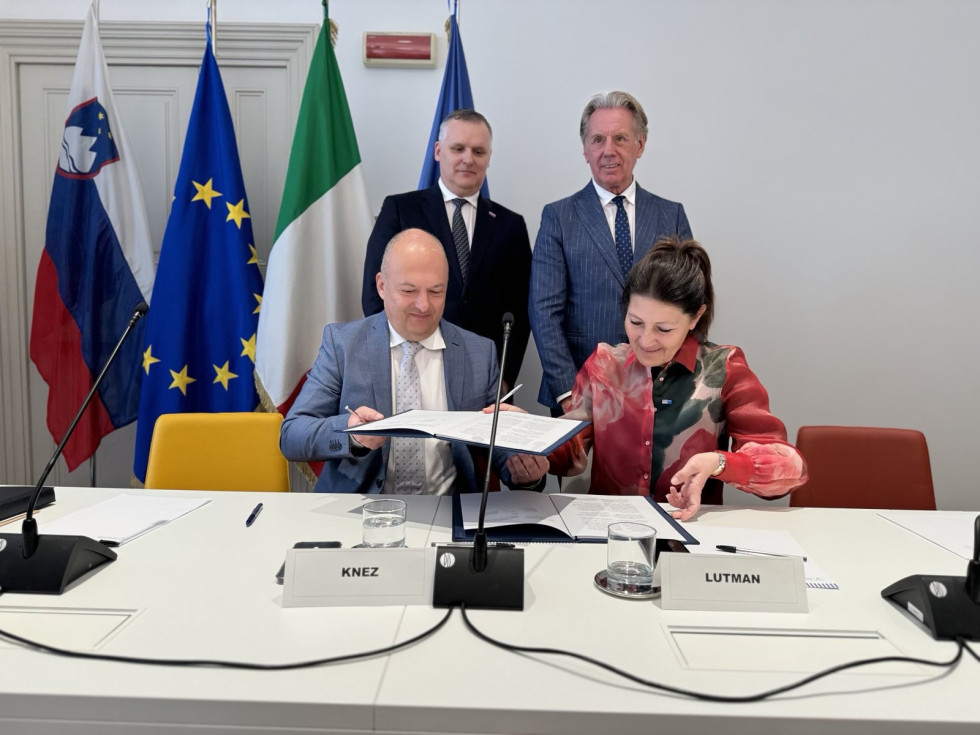 Podpisnika sedita za mizo in podpisujeta memorandum. Minister Kumer in Fabio Scoccimarro stojita v ozadju. Za njima evropska, slovenska in italijanska zastava. 
