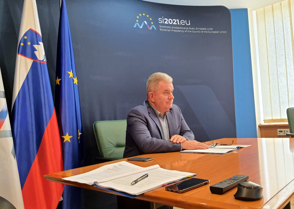 Minister Vizjak sedi za mizo in spremlja srečanje. Za njim je ozadje slovenskega predsedovanja ter slovenska in evropska zastava